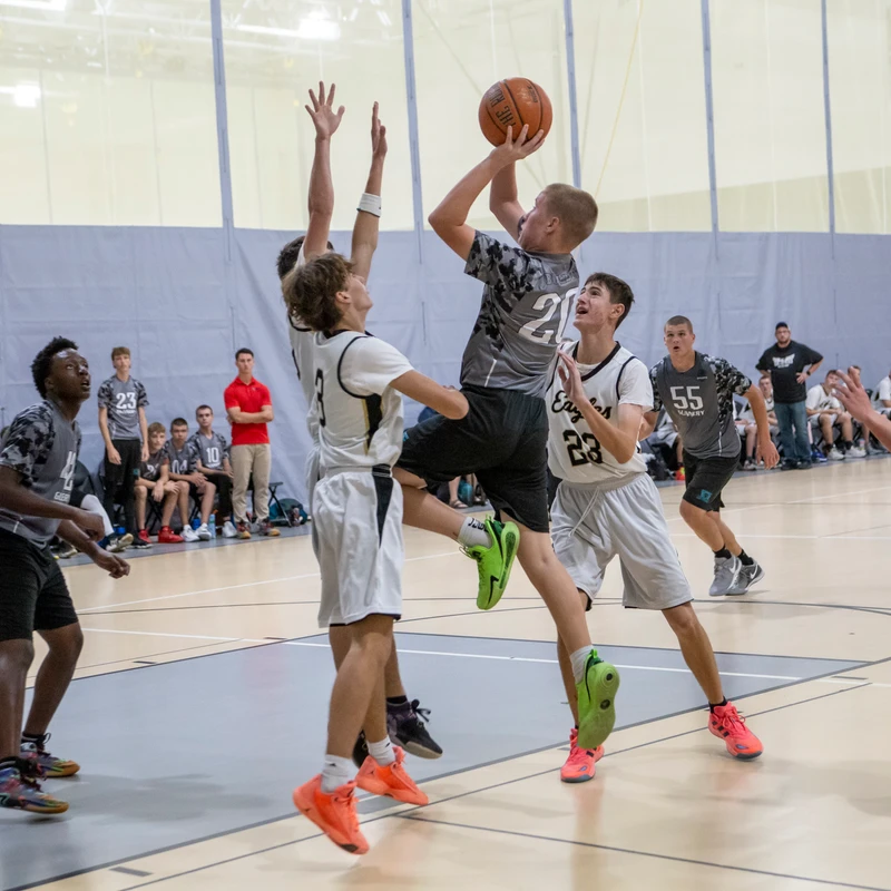 A young basketball player in a grey camo jersey jumps to shoot a layup while two players from the opposing team in white jerseys attempt to block the shot.