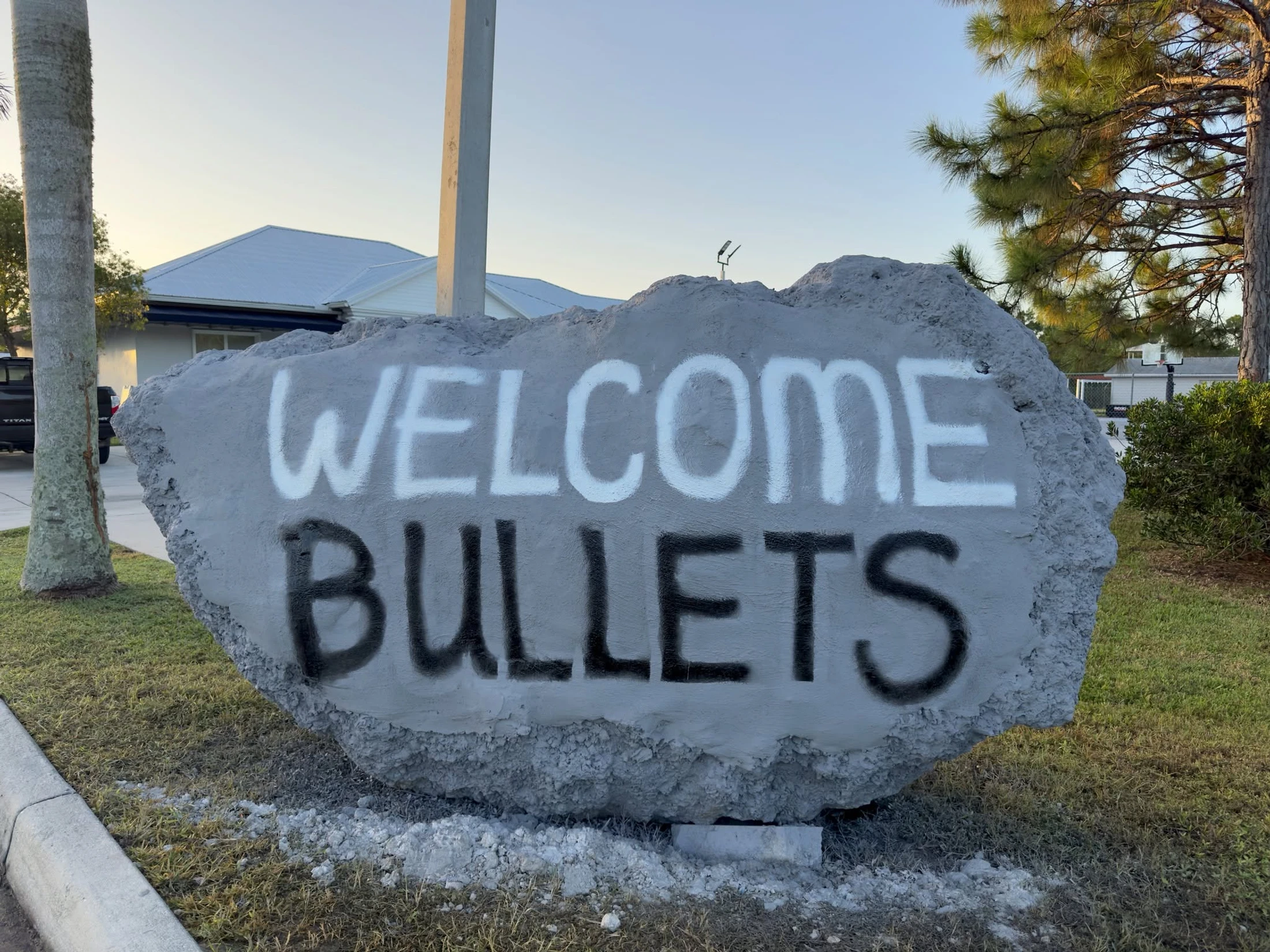 A large grey rock on a lawn spray-painted with the words 'WELCOME' in white and 'BULLETS' in black.