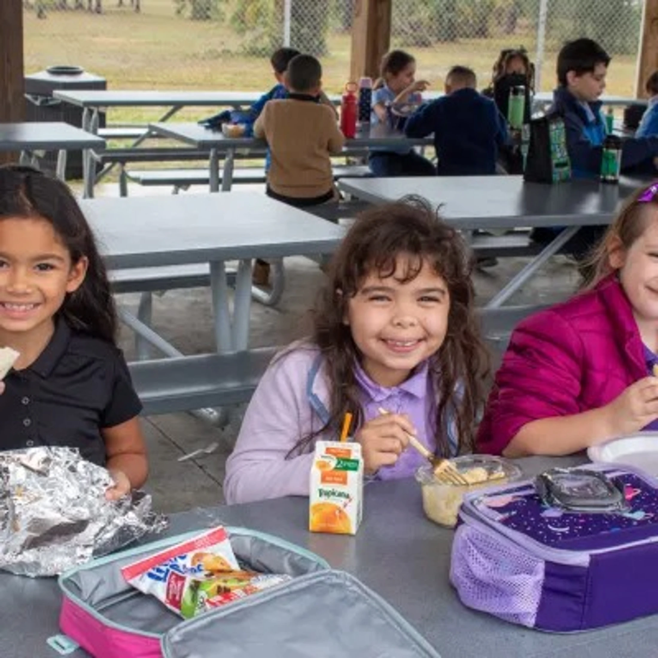 Three young girls smile for the camera while eating lunch with other students at outdoor picnic tables under a covered pavilion.