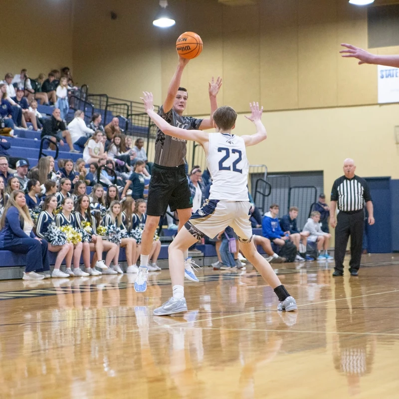 A male basketball player in a dark uniform jumps to shoot the ball as a defender in a white jersey with the number 22 attempts to block him in a gymnasium.