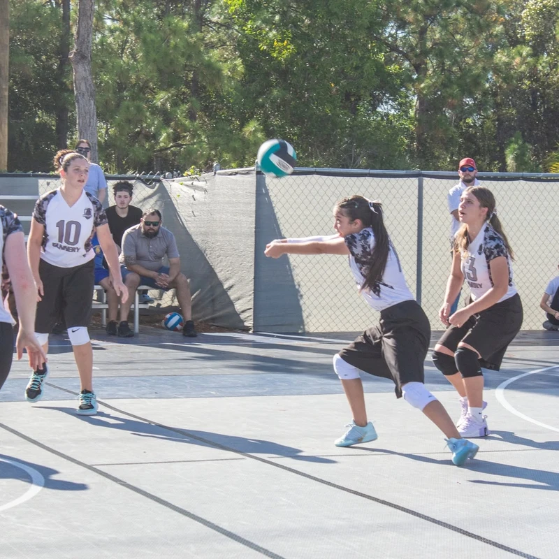 A young female volleyball player bumps the ball with her forearms during an outdoor game, with her teammates in position on the court and spectators watching.