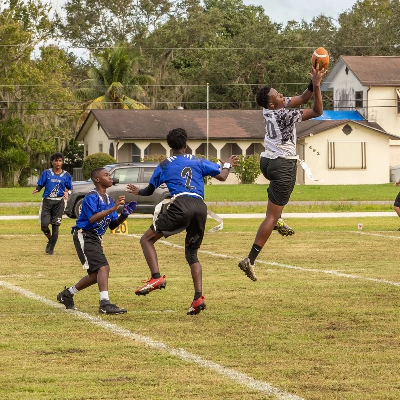 A young man leaps high in the air to catch a football during a flag football game, while other players in blue jerseys watch him on the grassy field.