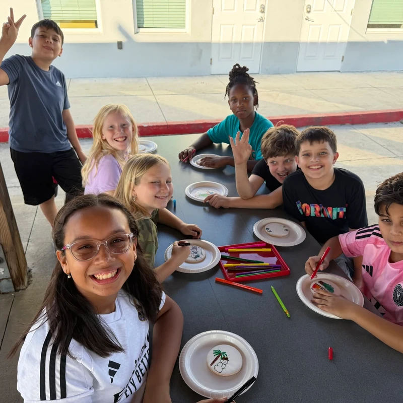 A diverse group of smiling children sits at an outdoor table, happily decorating round cookies with colorful markers and posing for the camera.