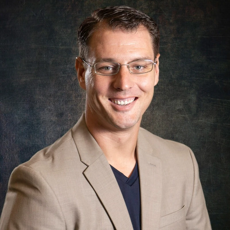 A headshot of a smiling man with short brown hair wearing glasses and a tan blazer over a blue shirt, set against a dark, textured background.