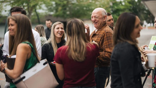 A group of people of various ages stand outdoors, talking and laughing together in a casual setting with trees in the background.