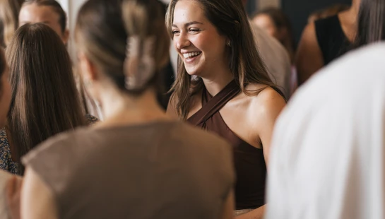 A young woman with long brown hair stands in a crowd, smiling widely as she talks to someone. The focus is on her happy expression.