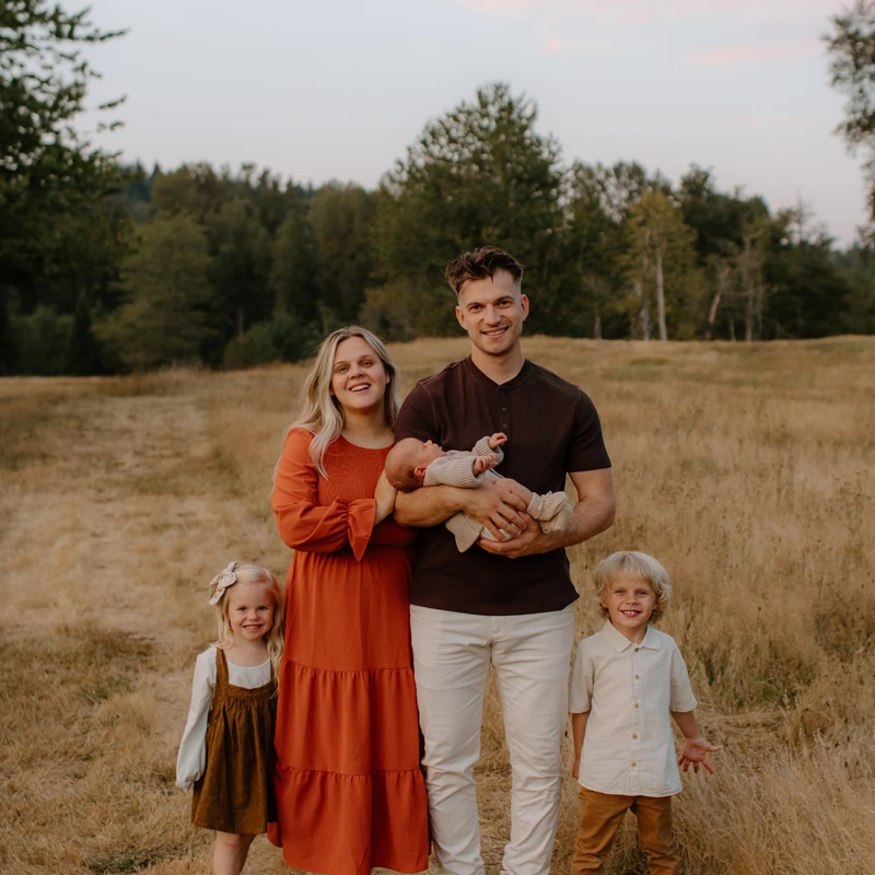 A smiling family of five, with a mother, a father holding a newborn, a young girl, and a boy, pose together in a golden, grassy field at dusk.