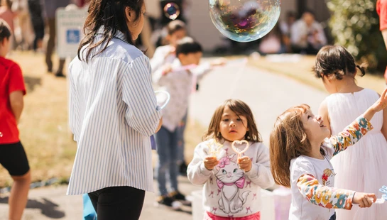 A group of children and adults outdoors on a sunny day. Young girls in the foreground look up in delight at large, floating soap bubbles.