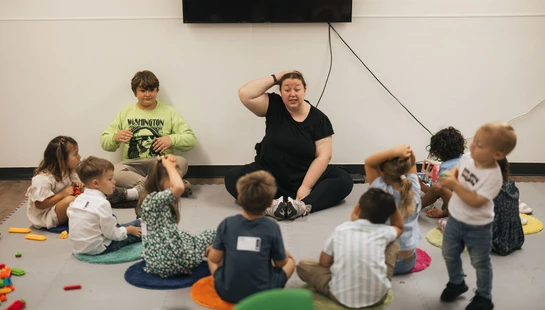 A female teacher sits on the floor in a circle with a group of young children, leading them in an activity where they touch their heads.