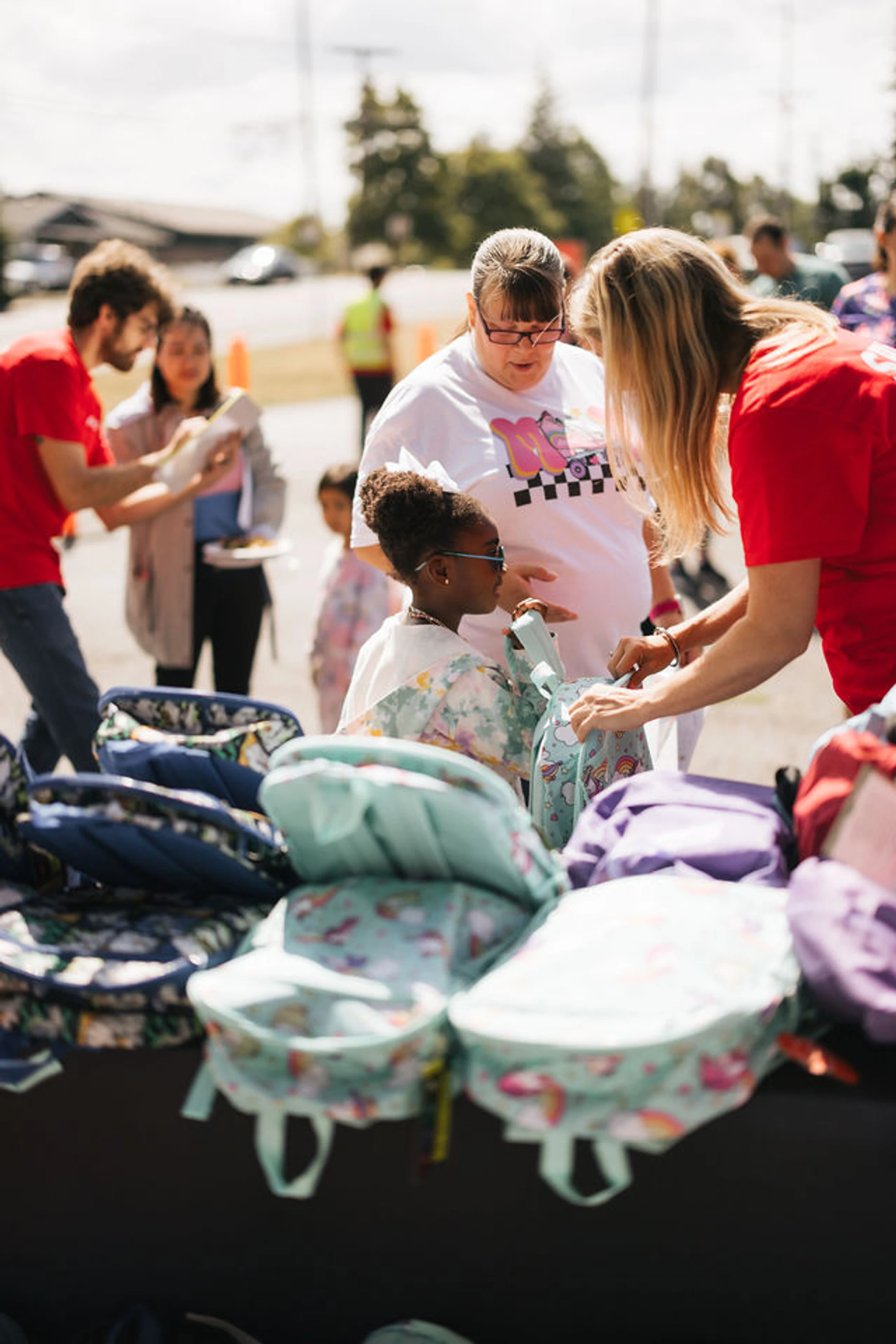 A young girl and her mother interact with a volunteer in a red shirt who is helping them pick out a new backpack from a table full of school bags.