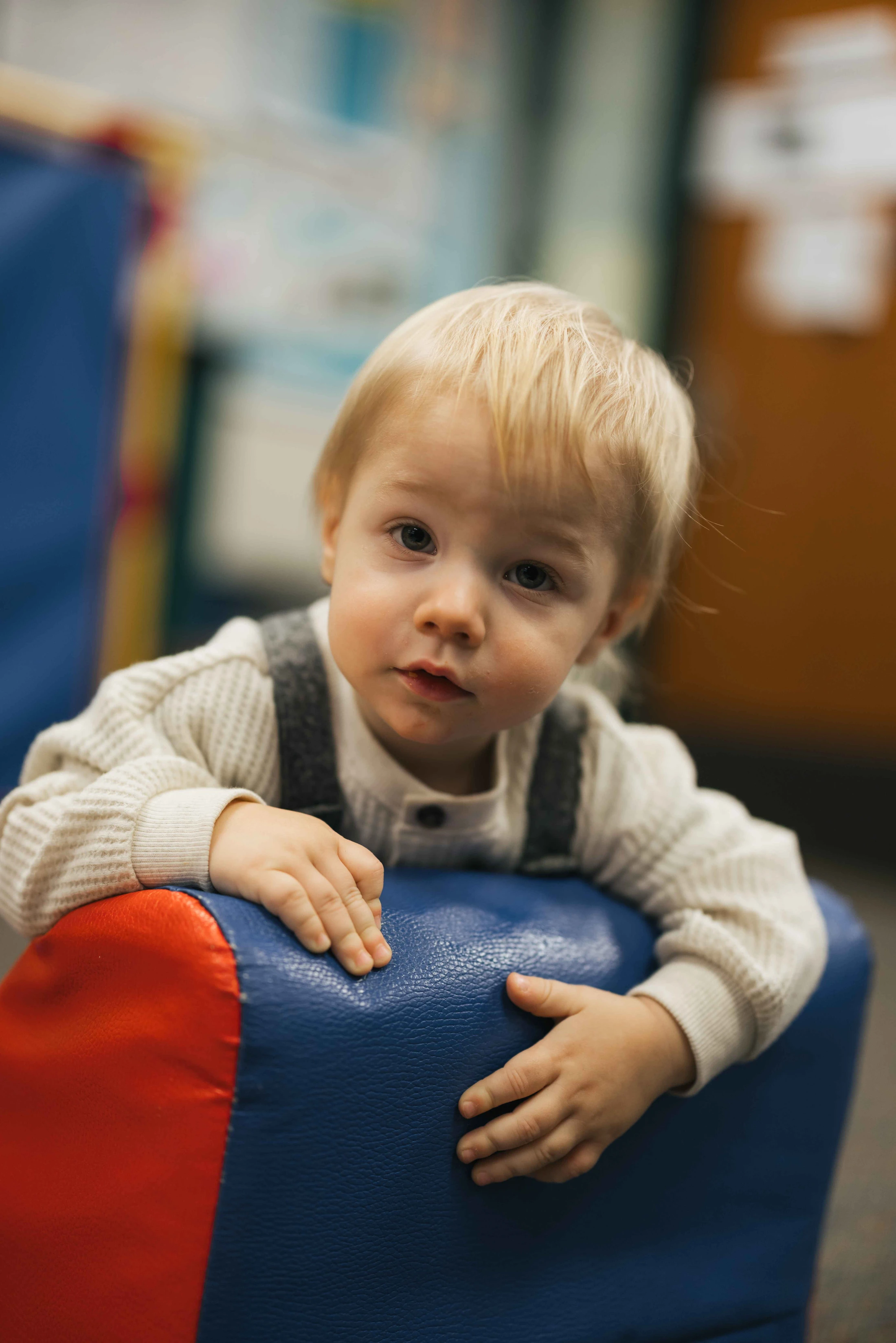 A close-up portrait of a toddler with blonde hair and light eyes looking at the camera while leaning on a blue and red piece of soft play equipment.