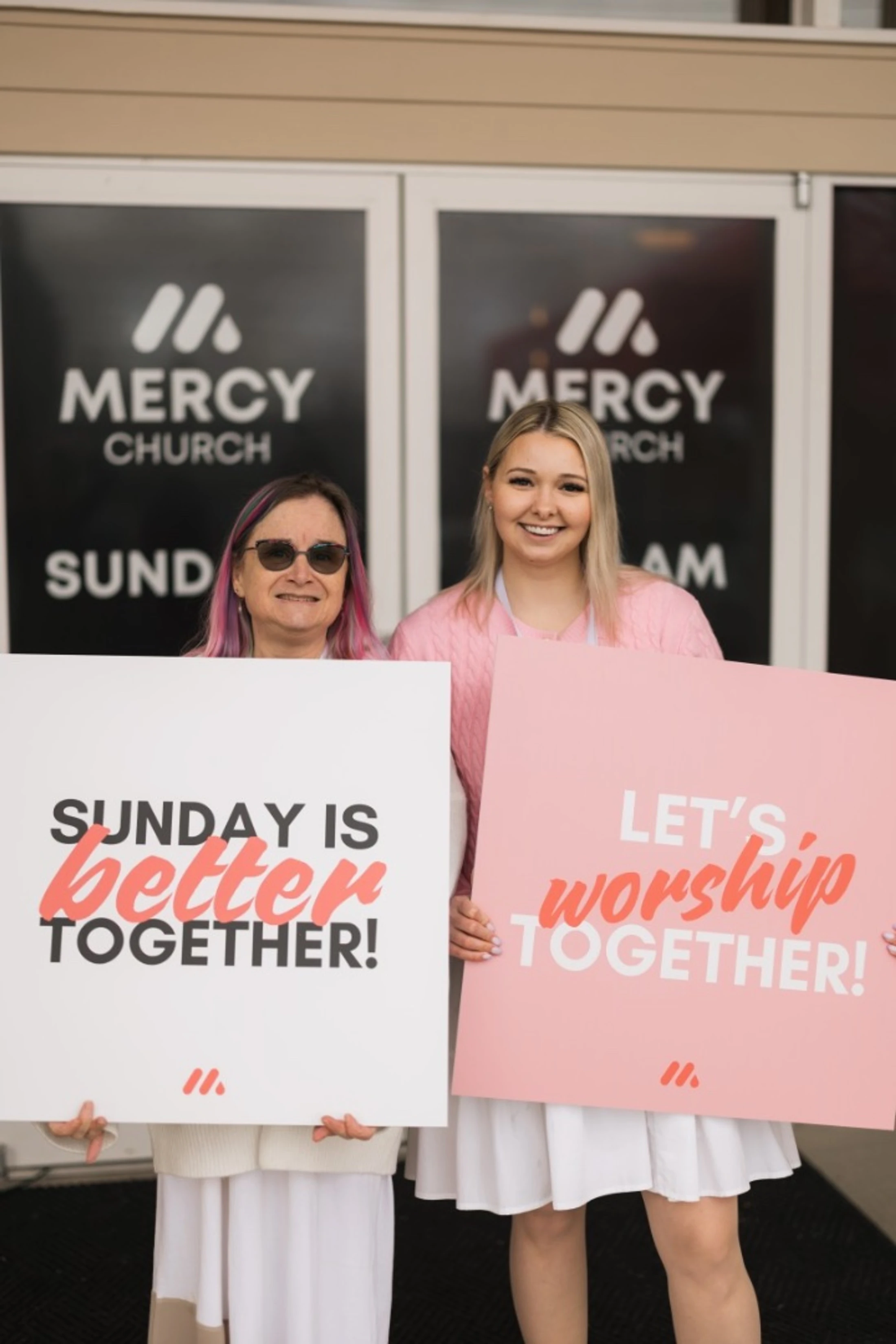Two women stand outside Mercy Church holding signs that read 'Sunday is better together!' and 'Let's worship together!' inviting people inside.