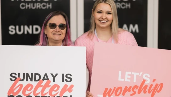 Two women stand outside Mercy Church holding signs that read 'Sunday is better together!' and 'Let's worship together!' inviting people inside.