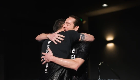 Two men in wet clothes hugging tightly inside a baptismal pool. The man on the right smiles with joy, his eyes closed, after being baptized.