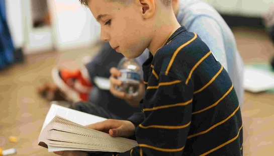 A young boy with a modern haircut sits cross-legged on the floor, wearing a striped shirt and intently reading a book he is holding.
