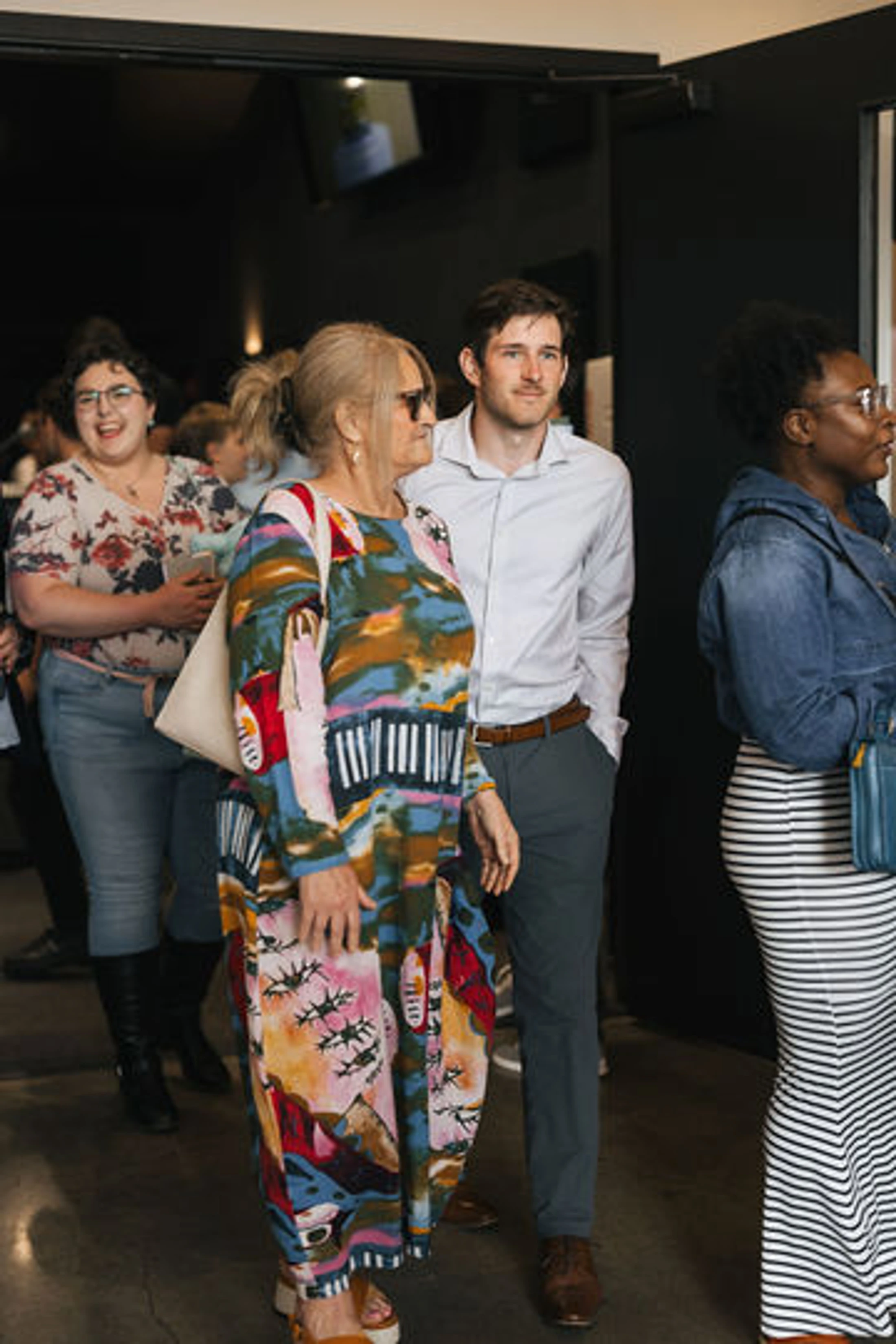 A diverse group of four people standing in a line indoors. A woman in a brightly colored patterned dress is in the center next to a man in a dress shirt.