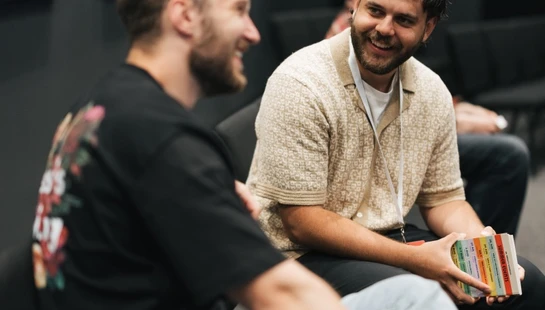 Two men sit talking in a dimly lit room. The man in focus smiles warmly, holding a small colorful book, while the other man in a black t-shirt smiles back.