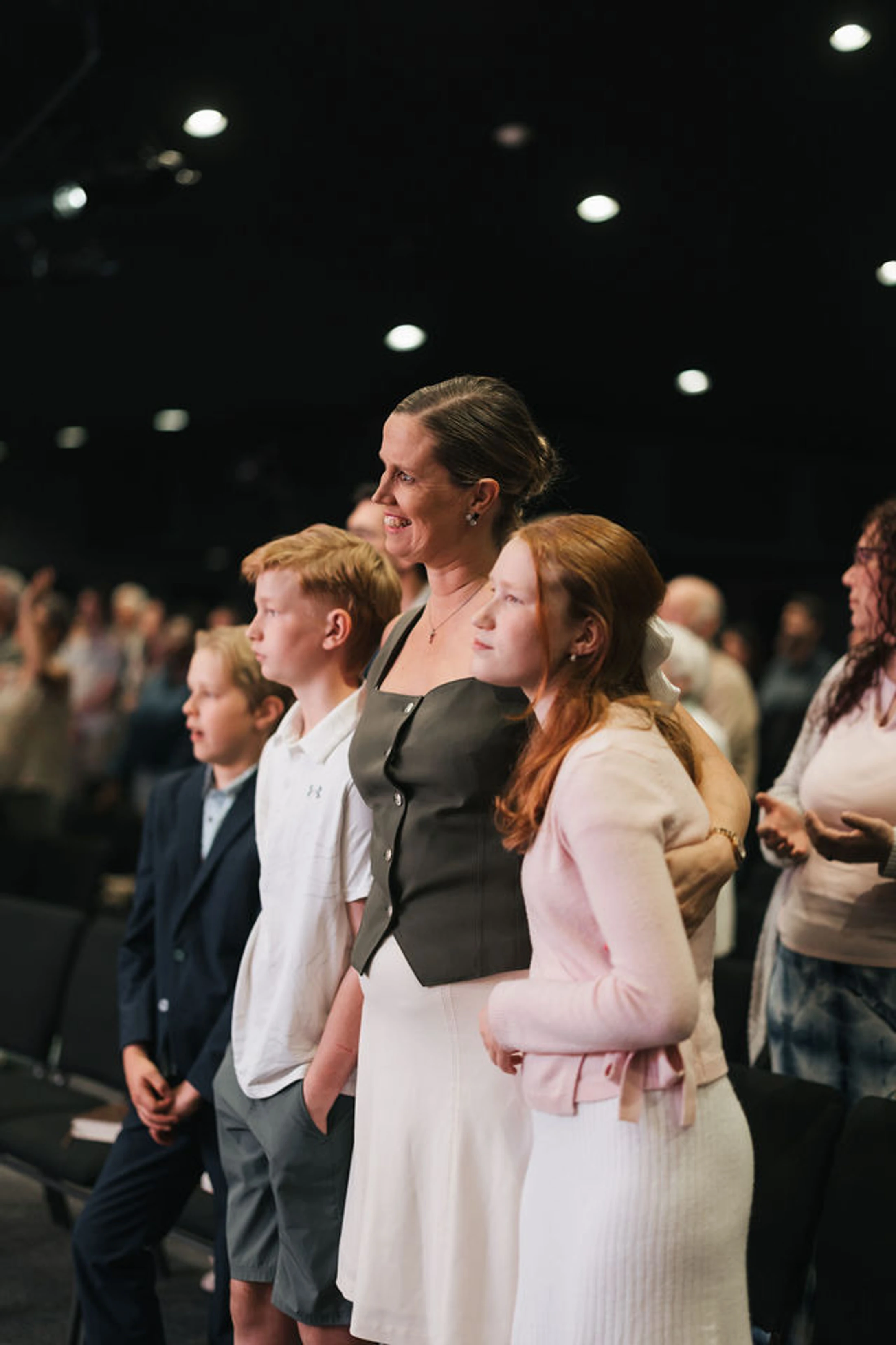A mother stands with her arm around her daughter, next to her two sons. They are in an audience, looking attentively at something off-camera.