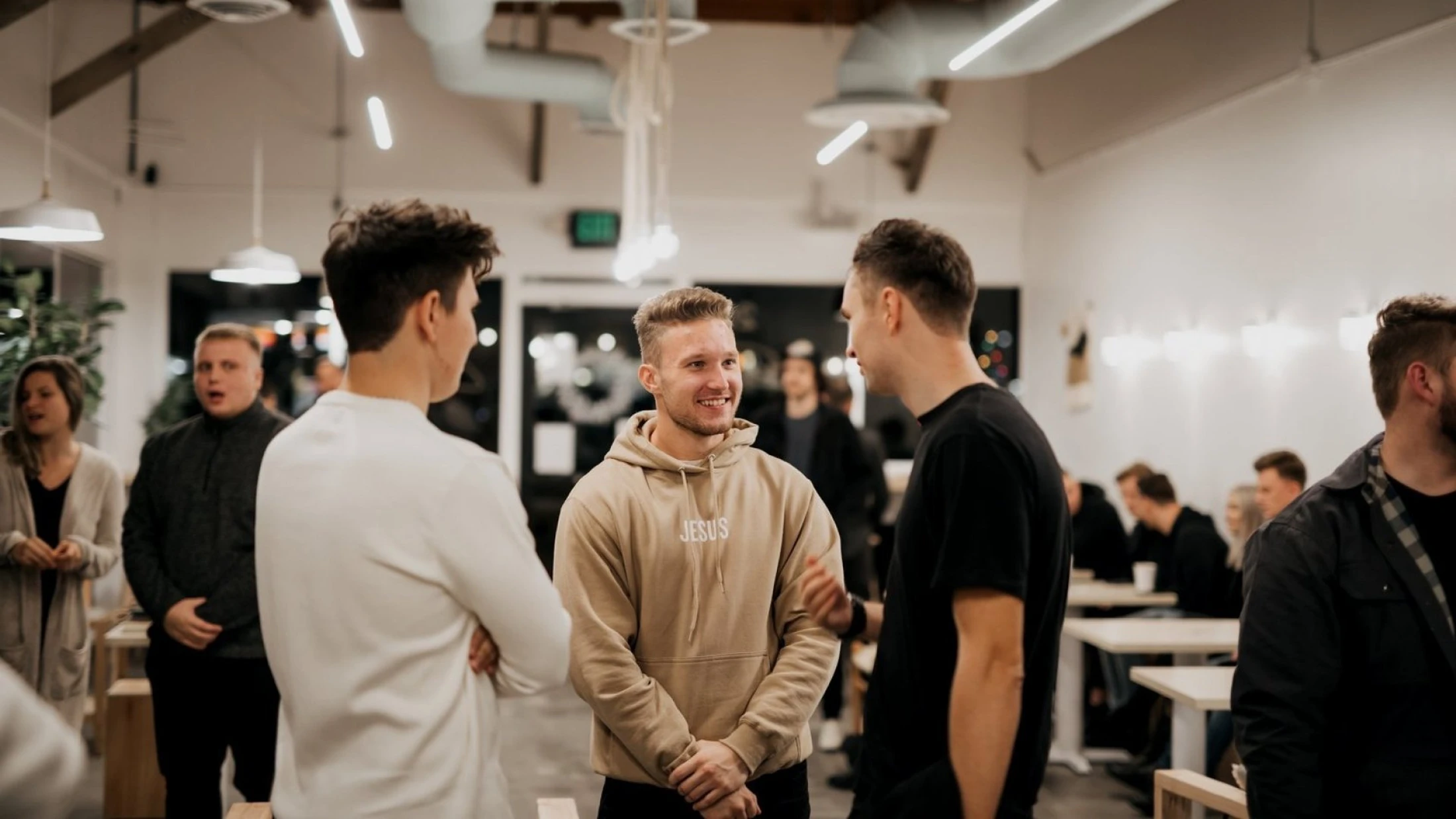 Three young men having a conversation in a well-lit room. The man in the center smiles warmly while wearing a beige hoodie with the word 'JESUS' on it.