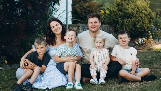 A smiling family of six, with a mother, father, and four young sons, sitting together on a green lawn in a garden for a portrait.