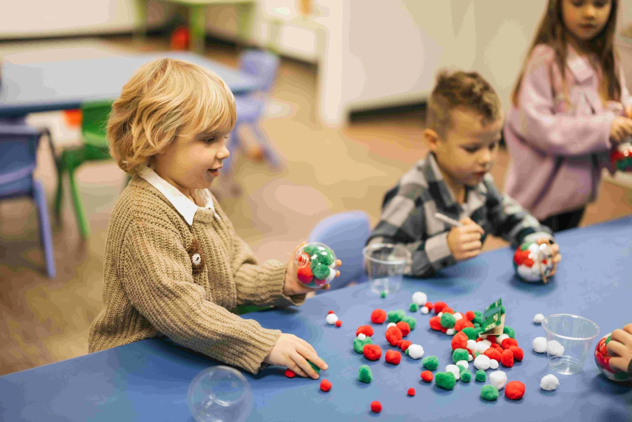 A young child with blonde hair smiles while filling a clear plastic ornament with red, green, and white pom-poms during a classroom craft activity.