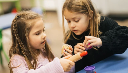 Two young girls sit at a blue table playing with modeling clay, a rolling pin, and a wooden tray full of metal cookie cutters in a bright classroom.