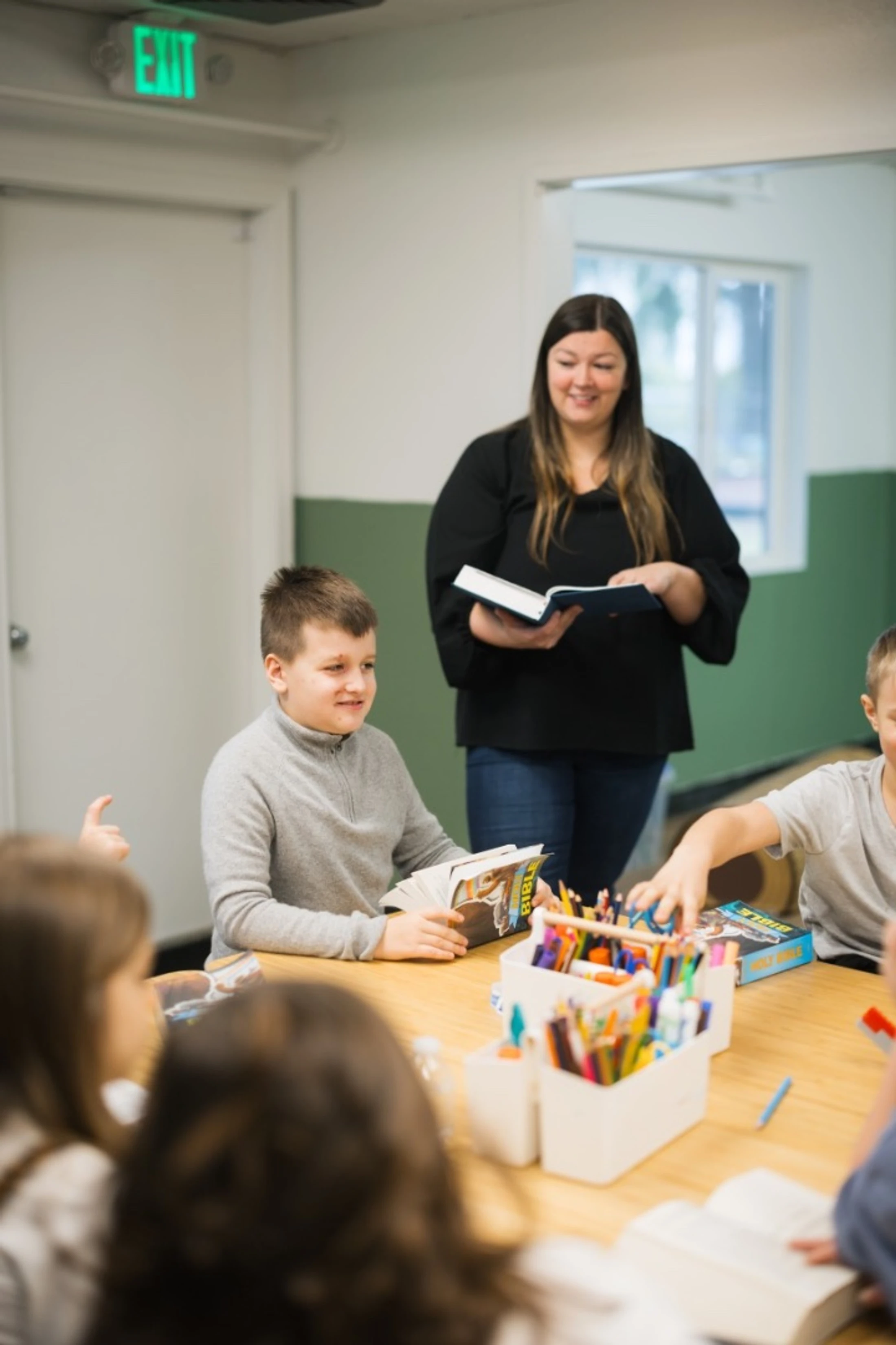 A female teacher smiles while holding a book, standing in front of a classroom of children sitting at a table with Bibles and art supplies.
