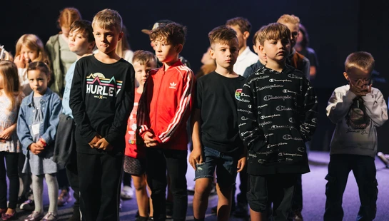 A group of young children, mostly boys, stand together on a dimly lit stage with a dark background, looking forward with serious expressions.
