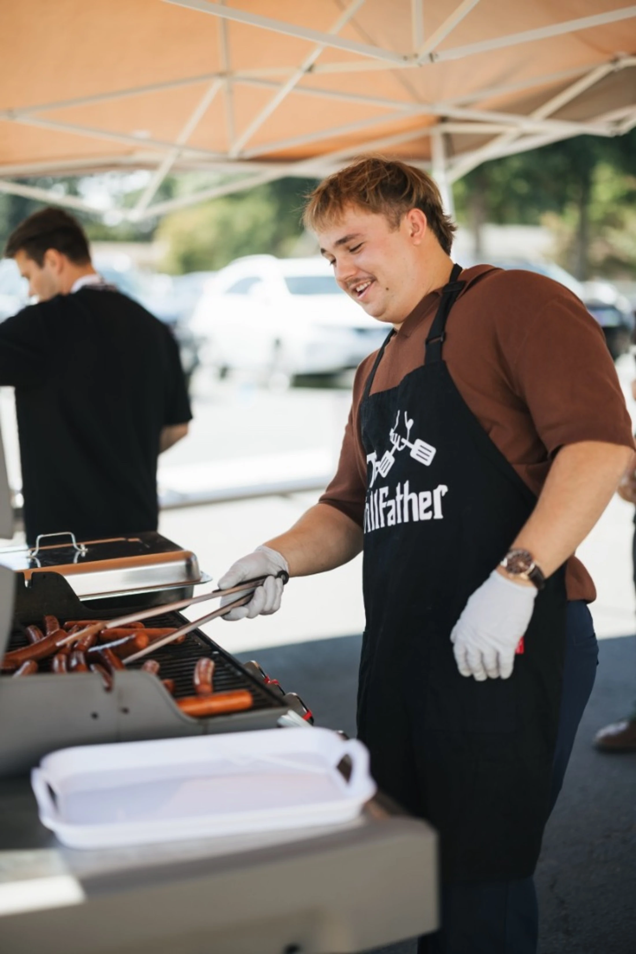 A smiling young man wearing a 'GrillFather' apron and disposable gloves uses tongs to cook hot dogs on a barbecue grill under a tent.