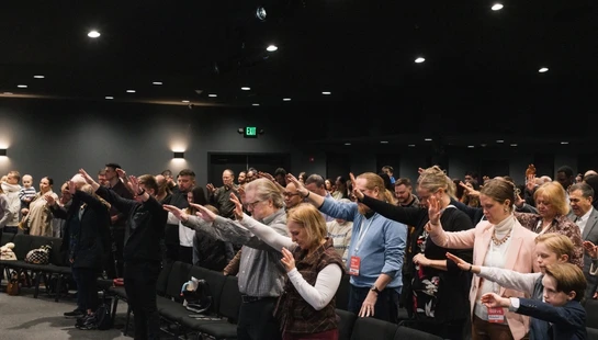 A diverse congregation stands between rows of chairs in a modern auditorium, raising their hands in a gesture of worship and prayer during a service.