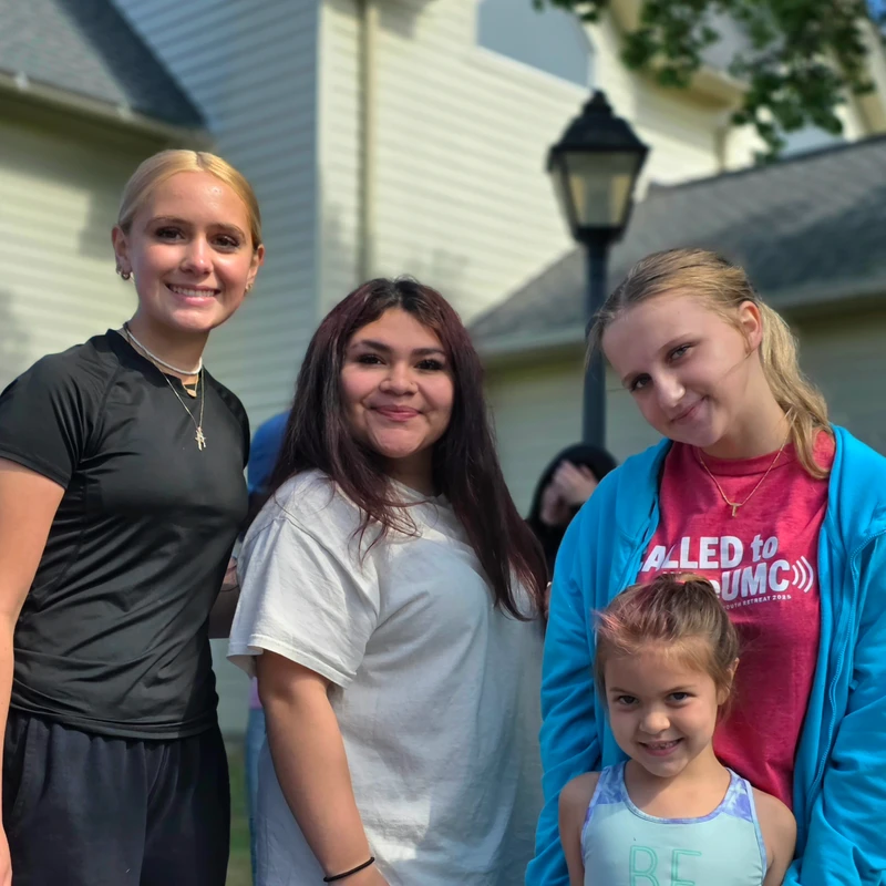 A group of four girls, including three teenagers and one young child, stand together outdoors and smile for the camera in front of a house.