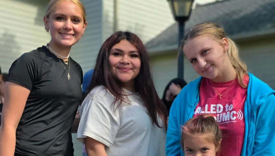 A group of four girls, including three teenagers and one young child, stand together outdoors and smile for the camera in front of a house.