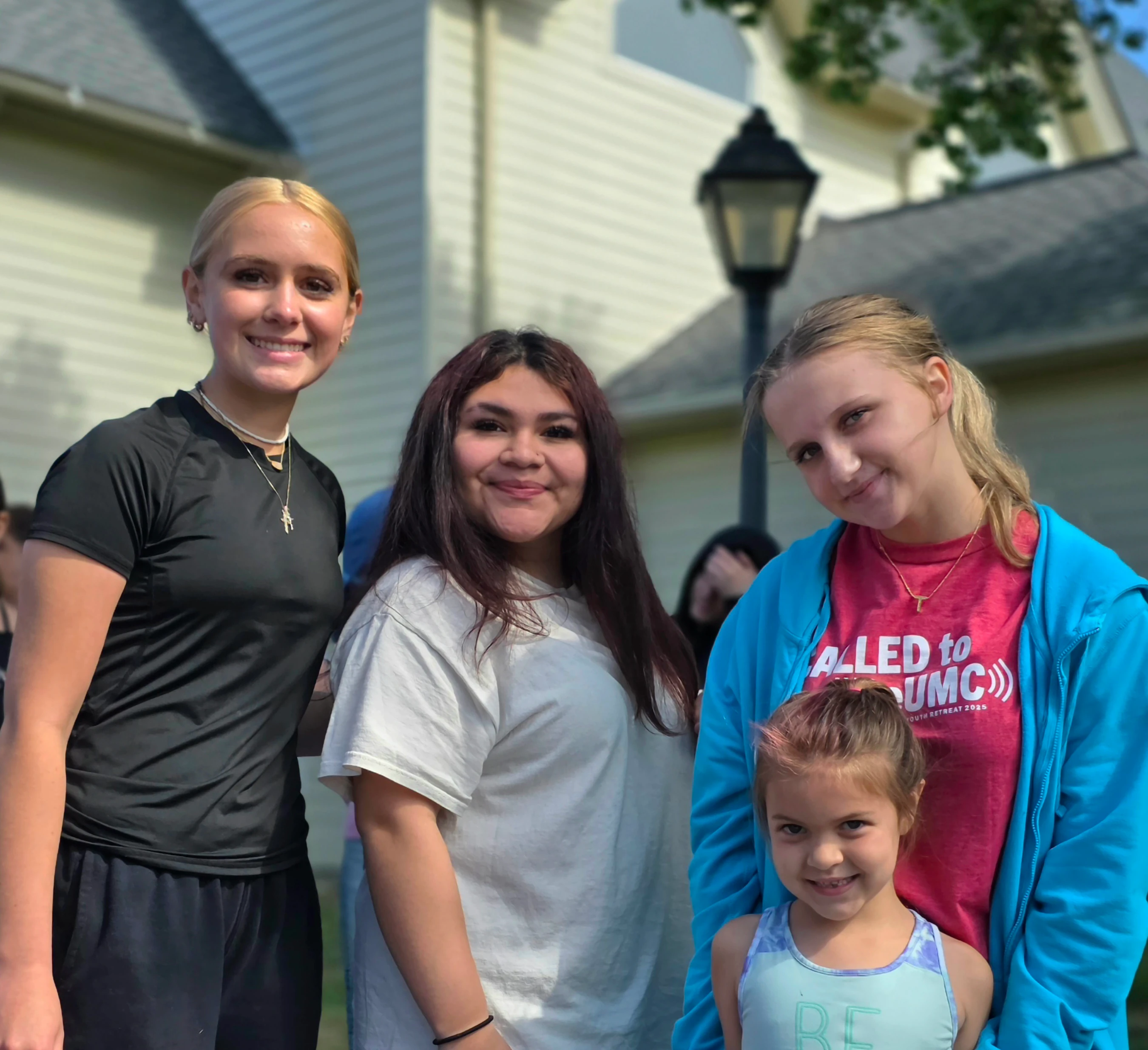 A group of four girls, including three teenagers and one young child, stand together outdoors and smile for the camera in front of a house.