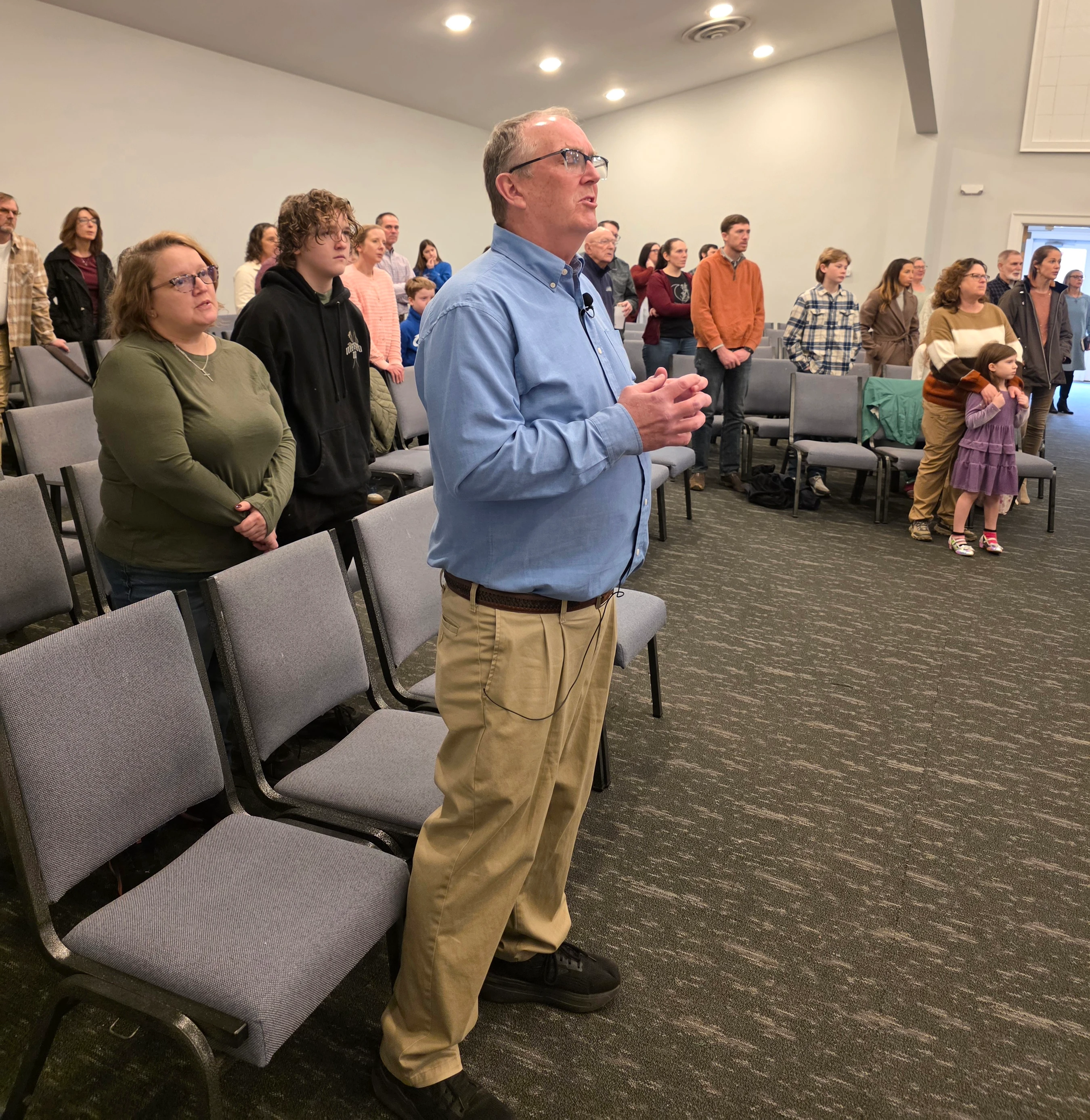 A diverse group of people standing in rows of chairs in a modern room, looking forward attentively. A man in a blue shirt and glasses is in the foreground.