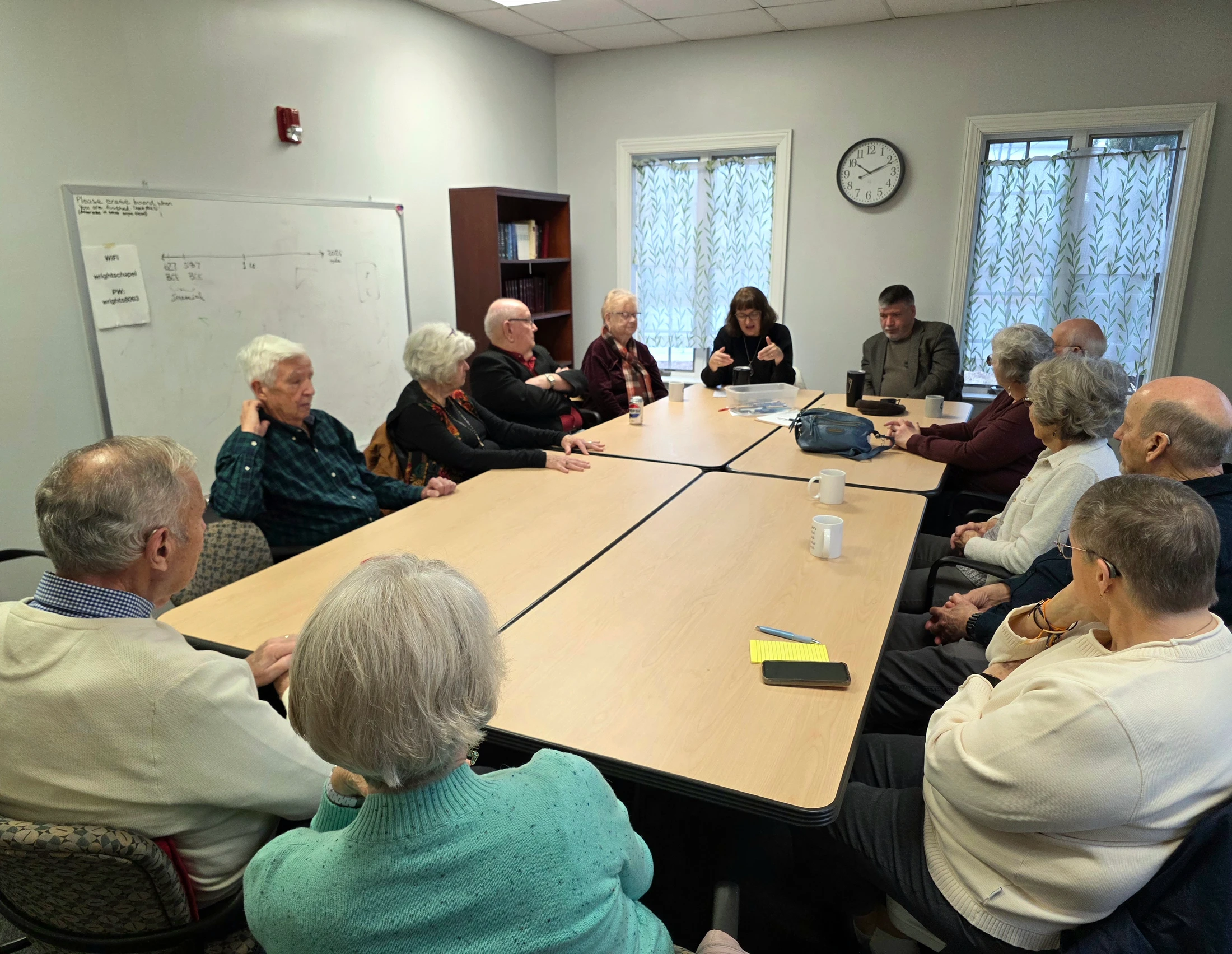 A group of older adults sit around a large conference table in a meeting room, engaged in a discussion led by a woman at the far end of the table.