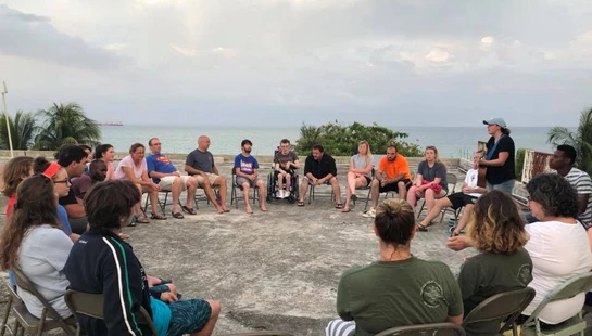 A diverse group of people sits in a circle on folding chairs on a concrete rooftop. In the background, the ocean stretches to the horizon under a cloudy sky.
