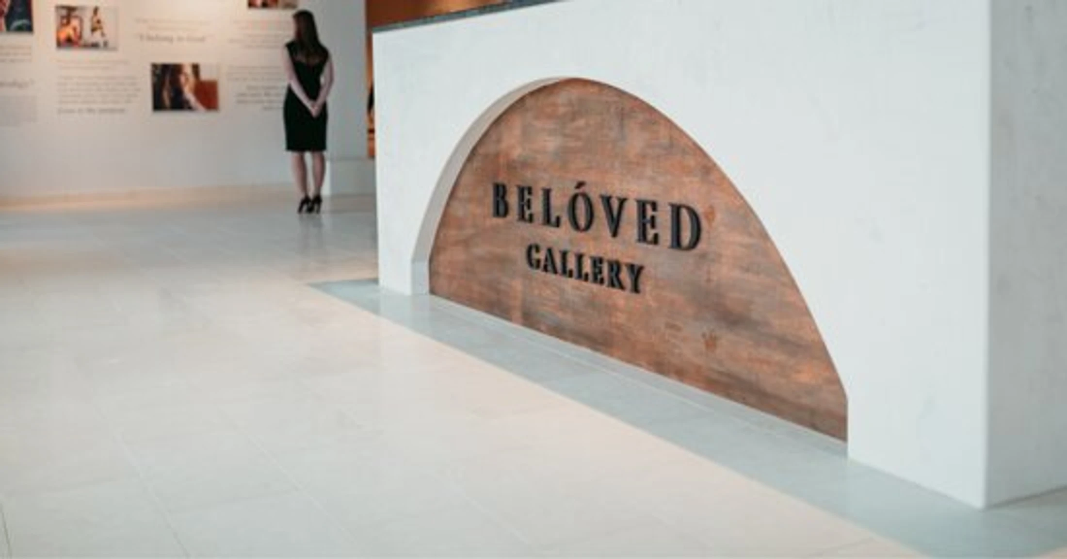 The interior of a modern art gallery with a sign for the Belóved Gallery on a white desk. In the background, a woman views an exhibition on the wall.
