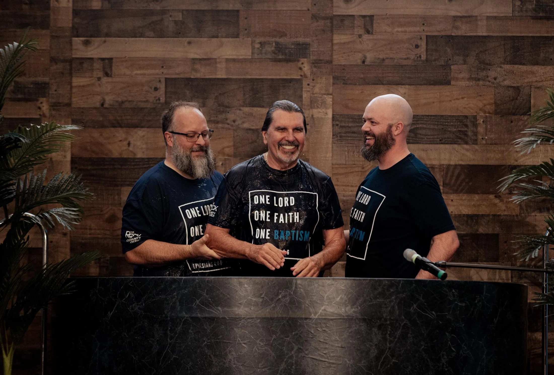 Three smiling men stand in a baptismal font. They are wet and wear matching black shirts reading "ONE LORD, ONE FAITH, ONE BAPTISM."