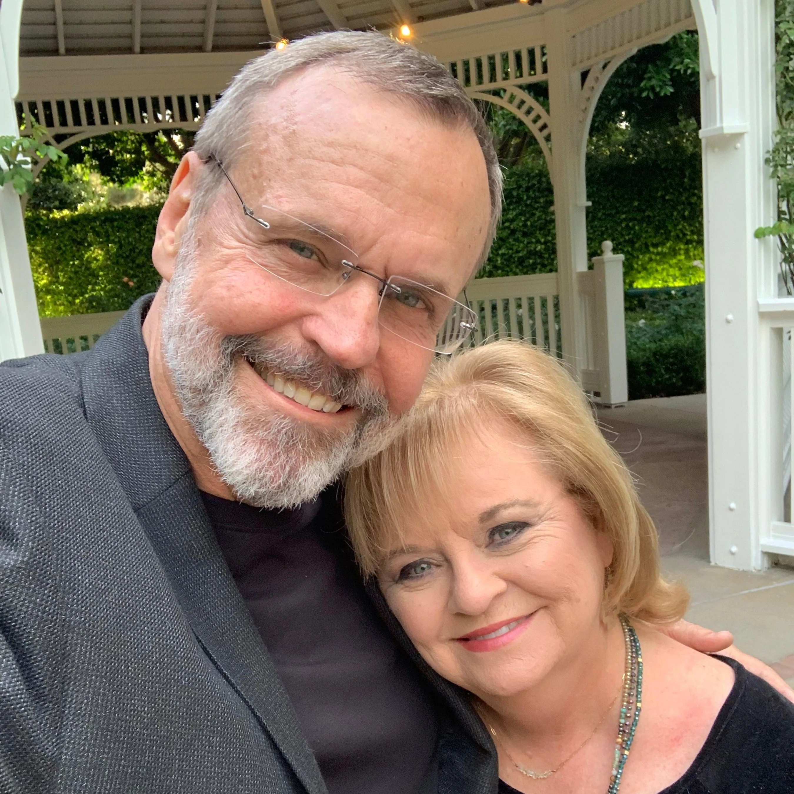 A smiling man with a gray beard and glasses takes a selfie with a blonde woman resting her head on his shoulder in front of a white gazebo.