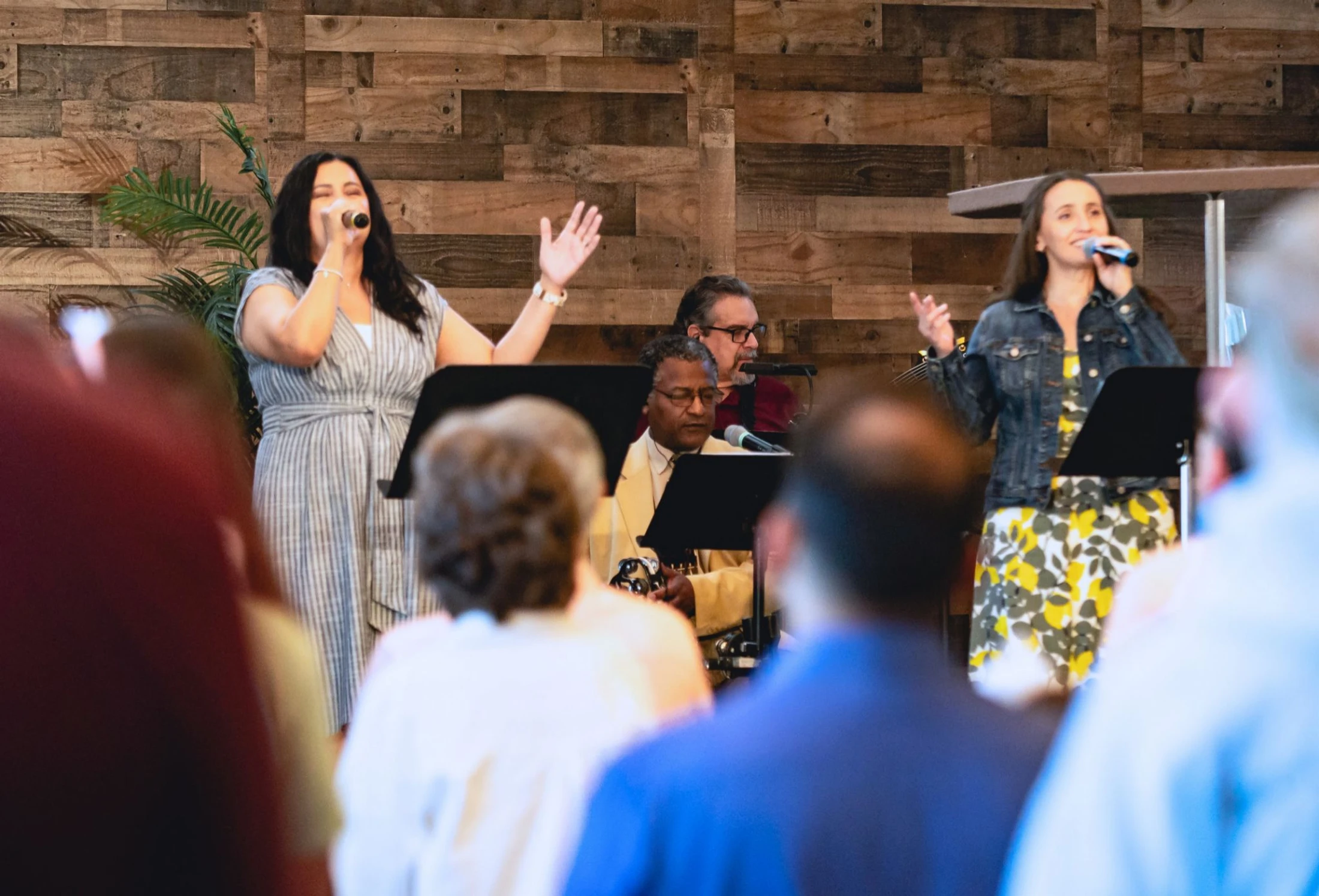 A diverse worship band performs on stage. Two female vocalists lead while male musicians play and sing behind them, in front of a rustic wood wall.