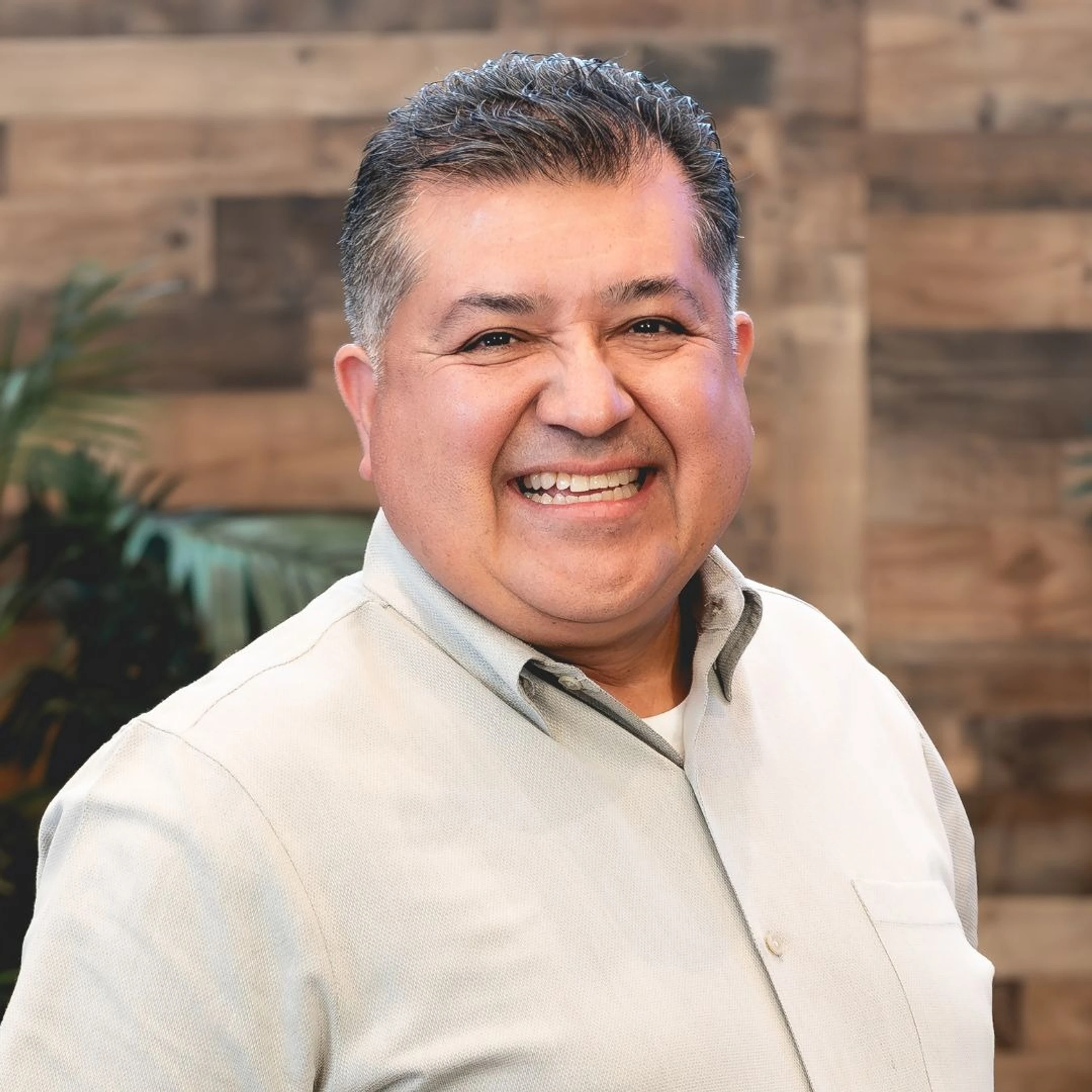 A headshot of a smiling, middle-aged Hispanic man with short dark hair, wearing a light gray button-down shirt against a blurred wooden background.