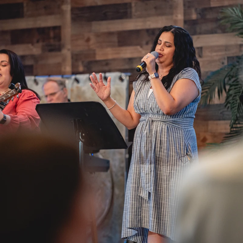 Two women lead music on a stage. One sings into a microphone with her hand raised, and the other sings while playing an acoustic guitar.