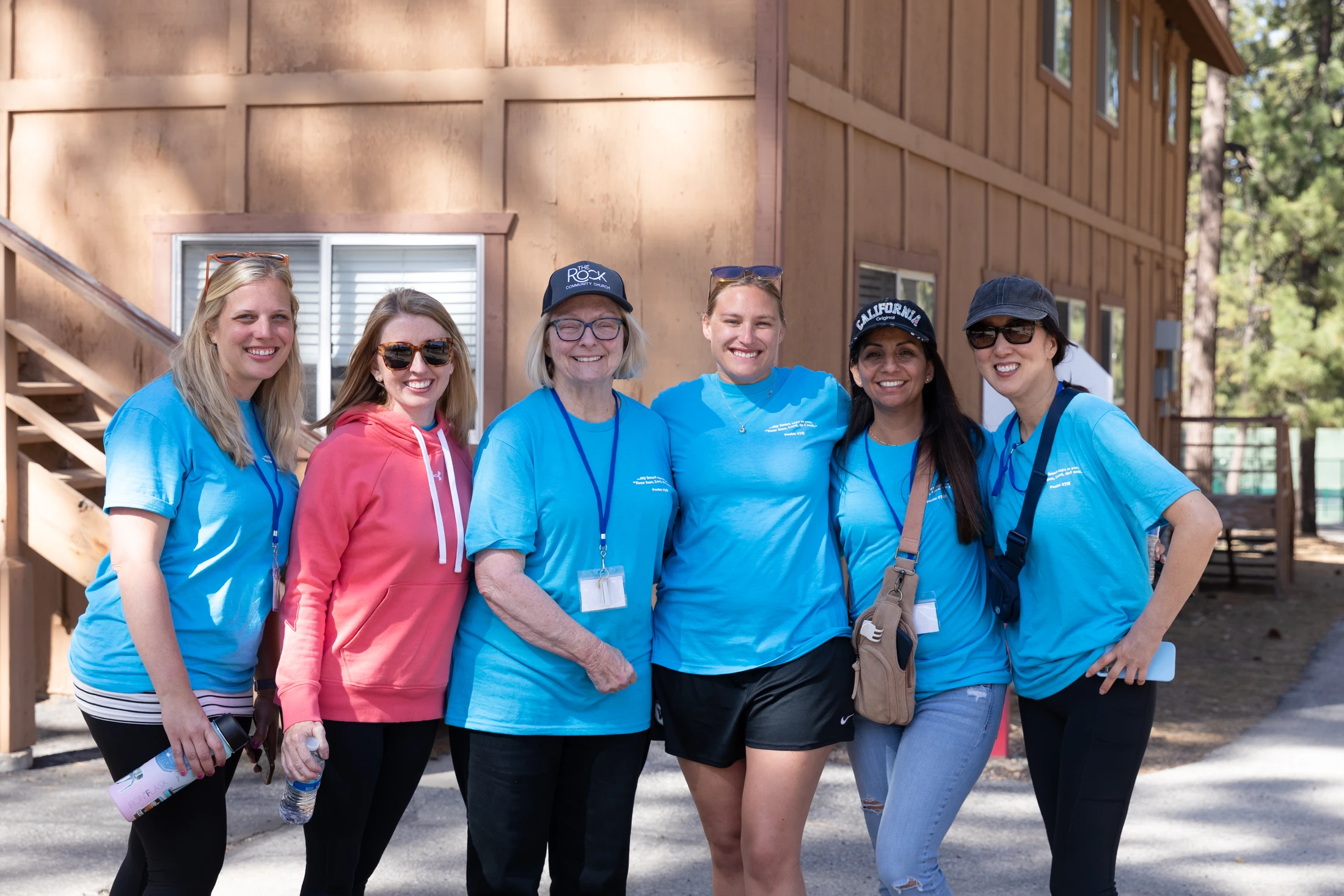 A group of six women in casual clothing smile for a photo outdoors in front of a wooden cabin. Most wear matching blue t-shirts.