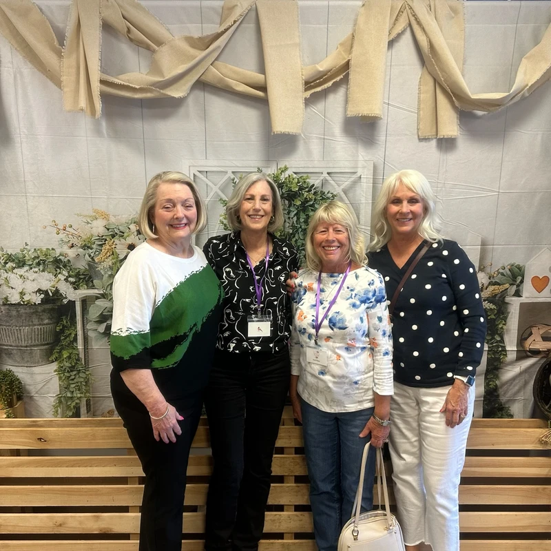 Four smiling women stand side-by-side for a group photo in front of a rustic backdrop decorated with greenery, wooden crates, and draped burlap.