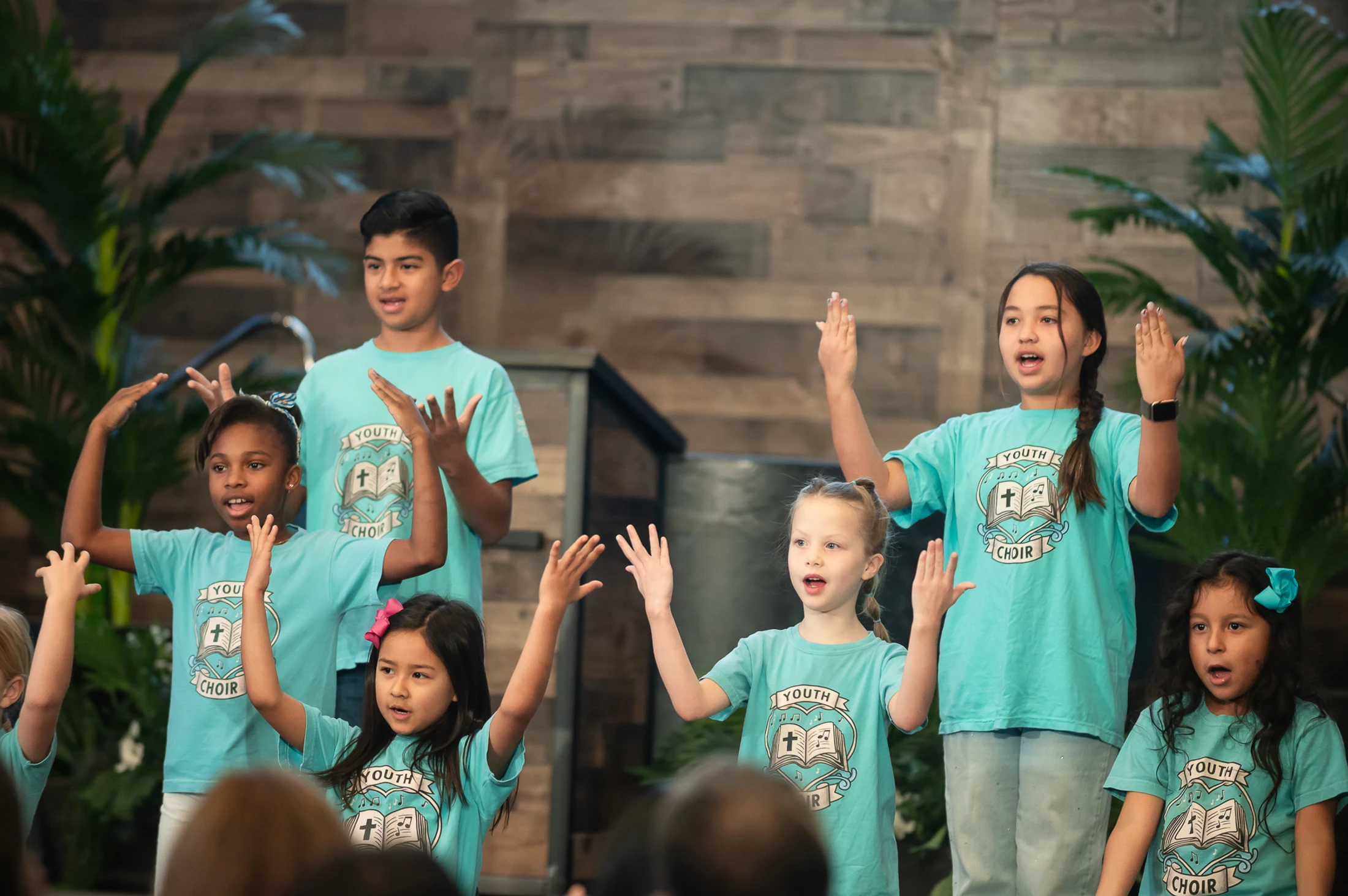 A diverse group of children in matching blue 'Youth Choir' t-shirts sing and use hand motions while performing together on a stage.