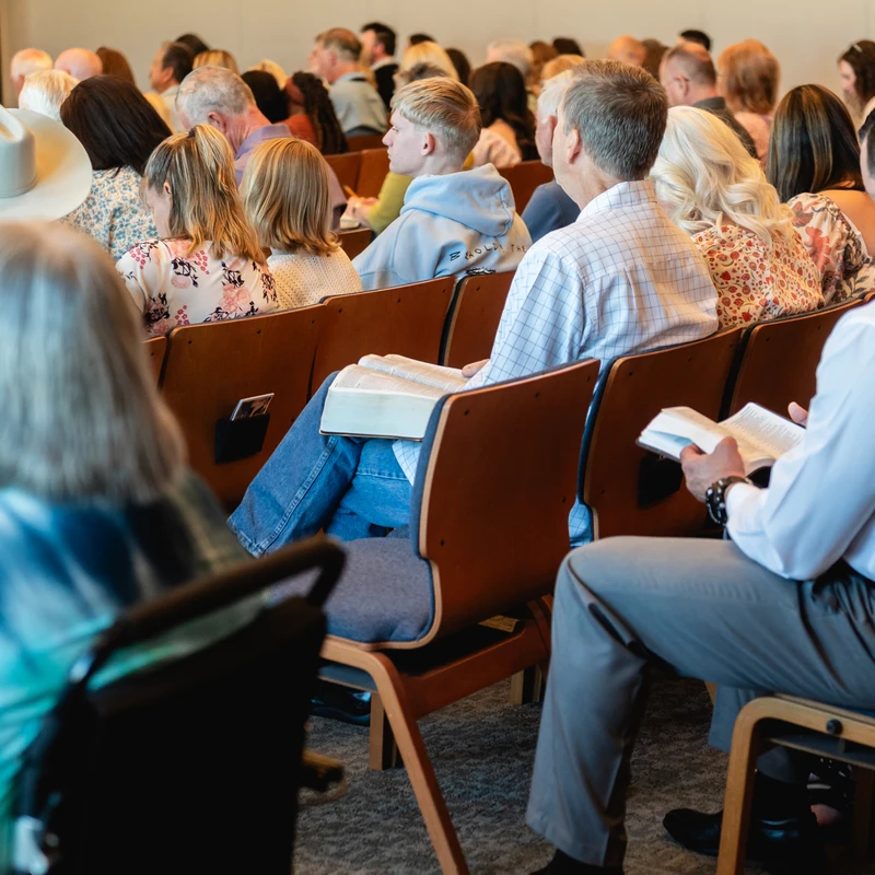 A diverse congregation sits in rows of wooden chairs, seen from behind. Many are listening intently, and some are reading from open books during a service.