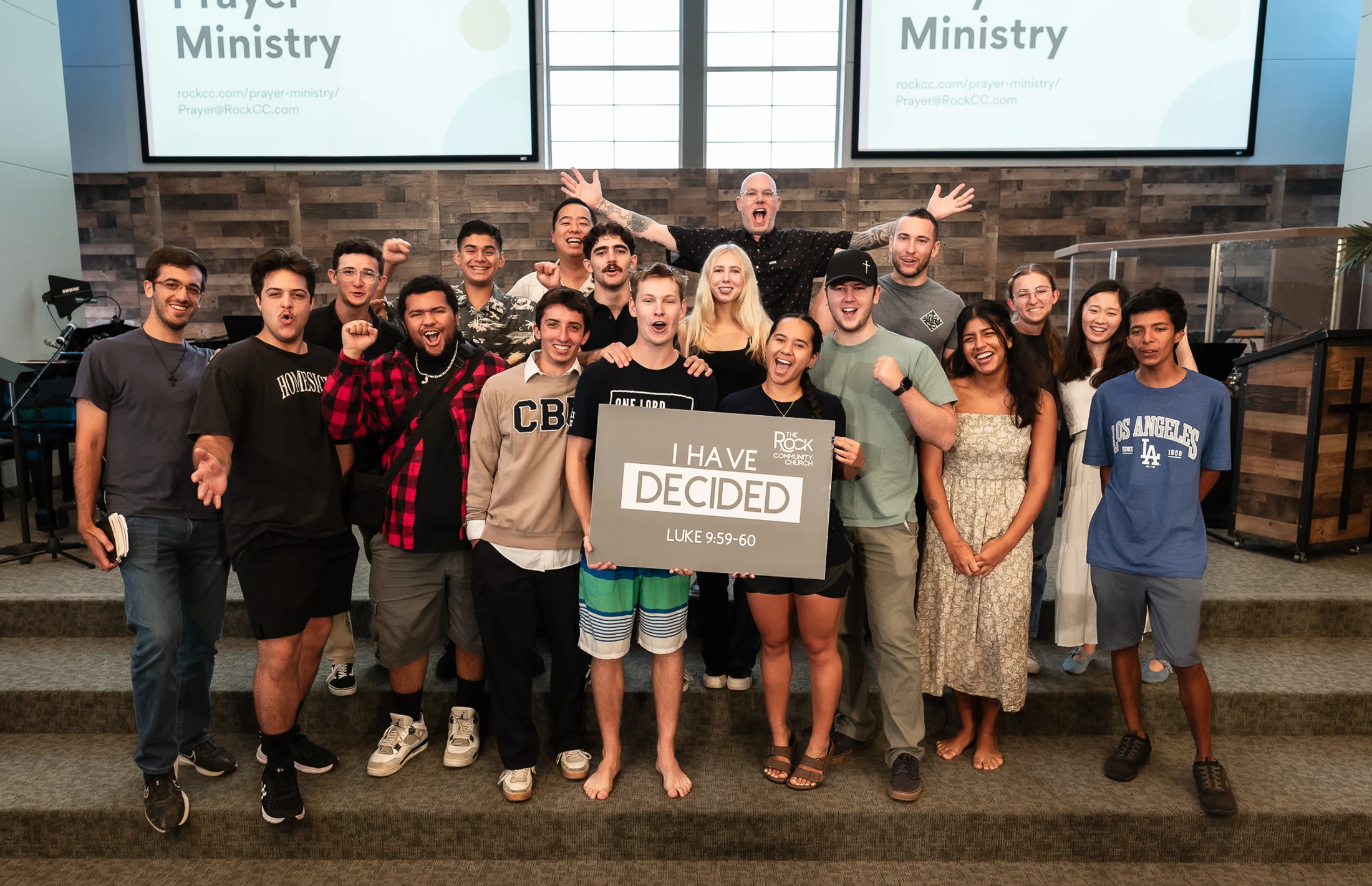 A diverse group of young adults and men pose for a photo on stage at a church. A man in the center holds a sign that says, 'I HAVE DECIDED.'