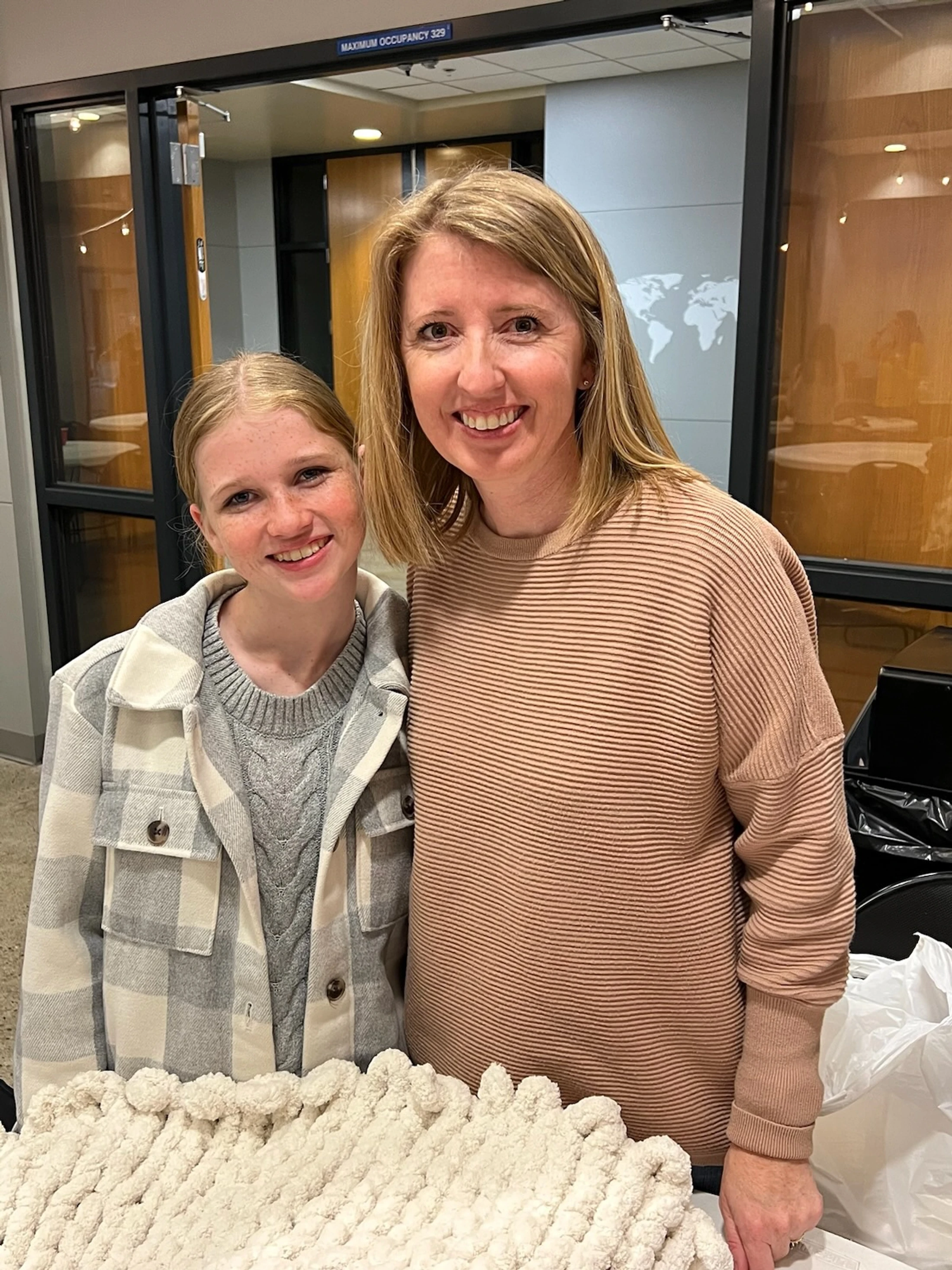 A young woman and an older woman, likely mother and daughter, smiling at the camera. In front of them is a large, white, chunky hand-knit blanket on a table.