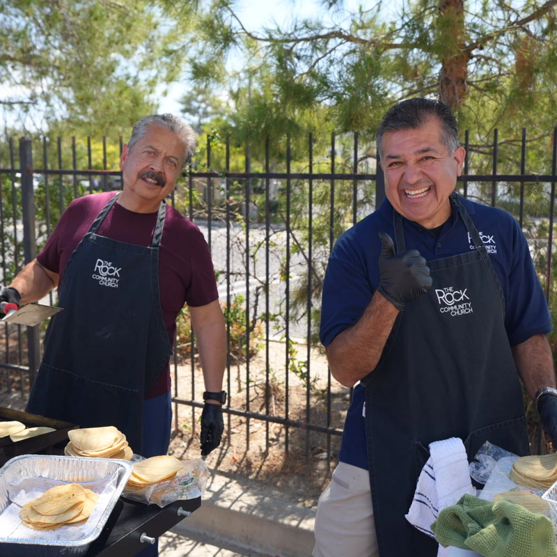 Two smiling men in church aprons cook corn tortillas on a large flat-top grill outdoors. One man gives a thumbs-up to the camera.