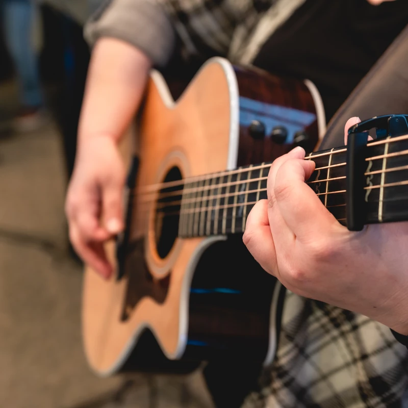 A close-up of a person's hands playing an acoustic guitar. One hand is fretting a chord near a capo clamped onto the neck of the instrument.